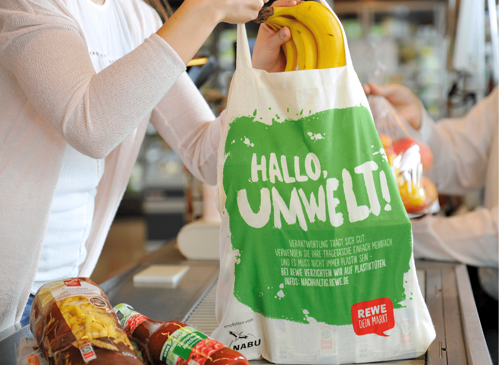 REWE checkout scene with employee handing groceries to a customer.