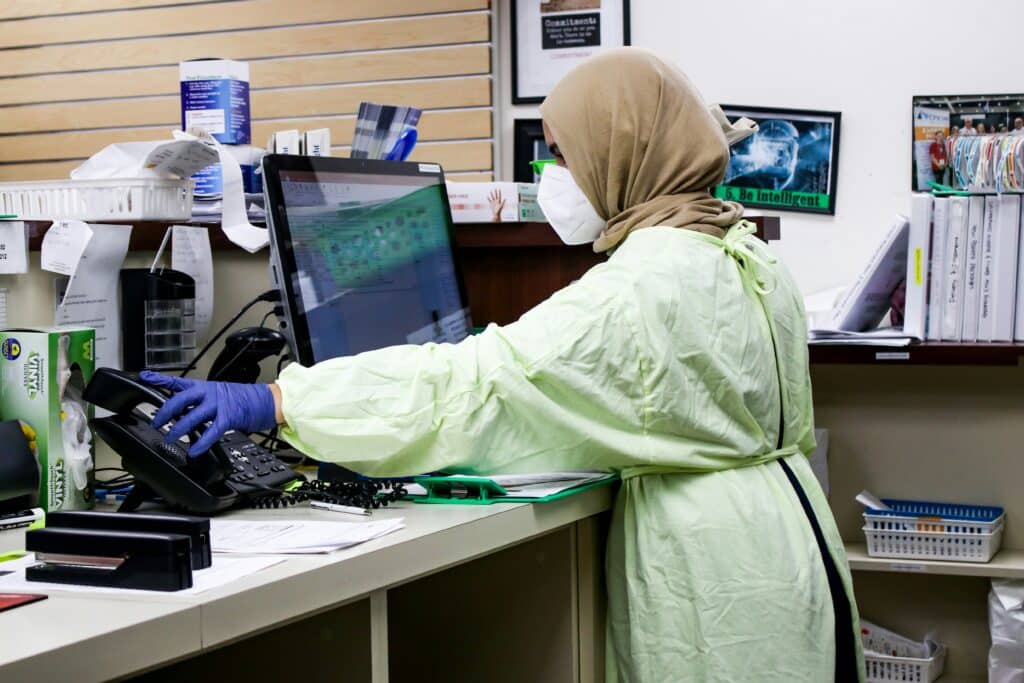 woman in green lab coat and purple gloves at her laptop and using a phone