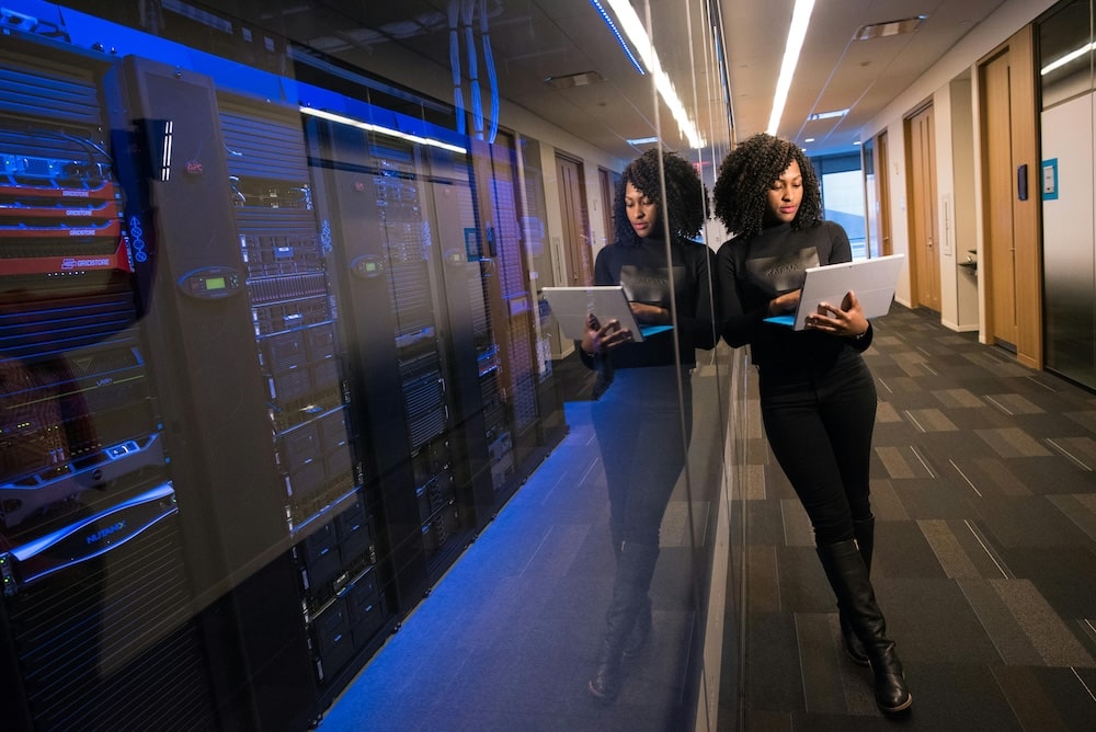 woman with curly hair, on her laptop wearing black, standing in a hall looking at market research data